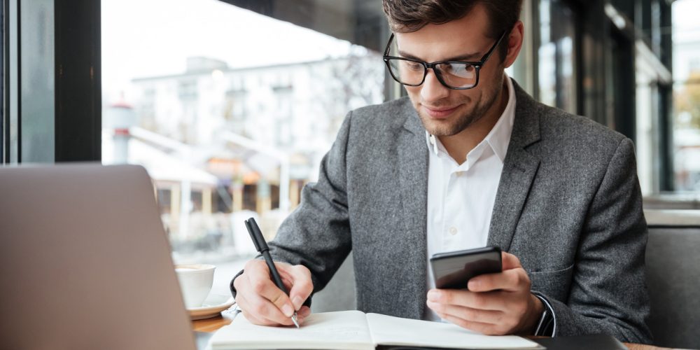 smiling-businessman-eyeglasses-sitting-by-table-cafe-with-laptop-computer-while-using-smartphone-writing-something_Easy-Resize.com
